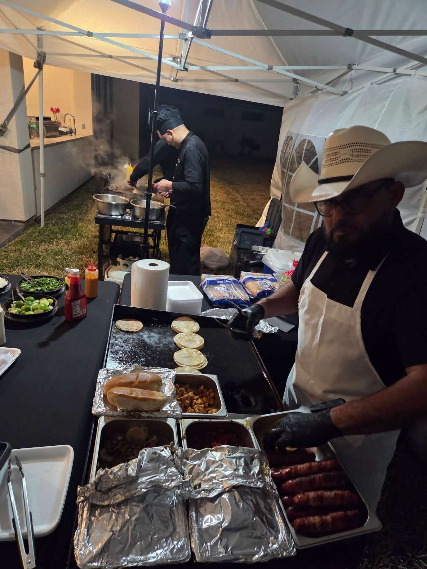 Taquero grilling tortillas and bacon-wrapped hot dogs under the tent